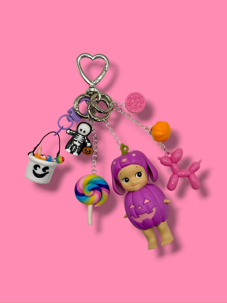 Purple Halloween Pumpkin Sonny Angel bag charm with skeleton, ghost candy bucket, pink Oreo, balloon dog and rainbow lollipop on pink background