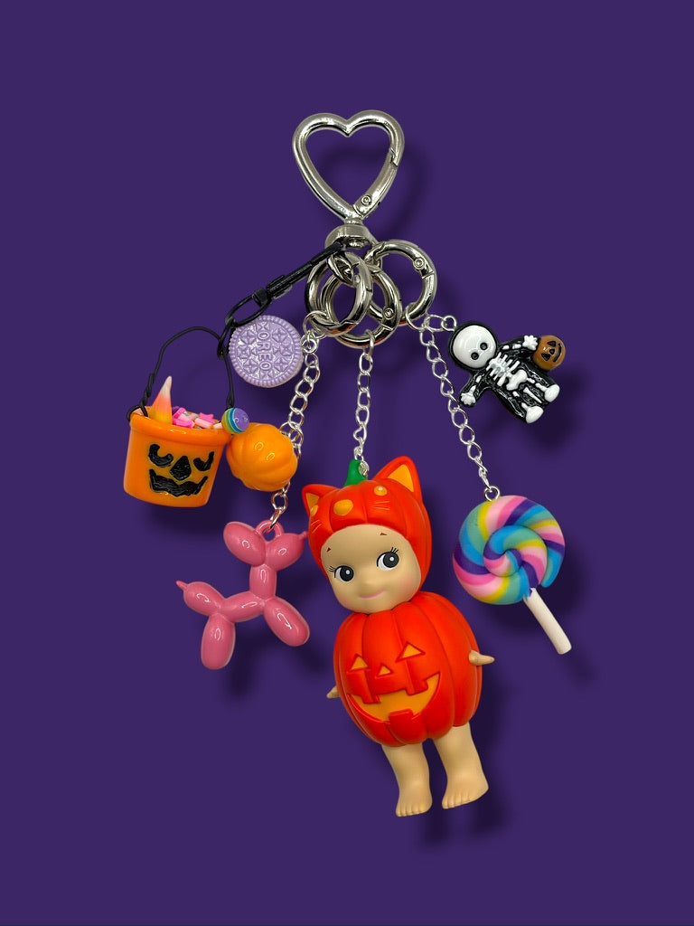 Halloween Pumpkin Sonny Angel bag charm with skeleton figure, jack-o-lantern bucket, Oreo charm, balloon dog, and rainbow lollipop on purple background