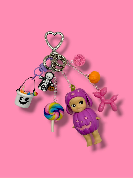 Purple Halloween Pumpkin Sonny Angel bag charm with skeleton, ghost candy bucket, pink Oreo, balloon dog and rainbow lollipop on pink background